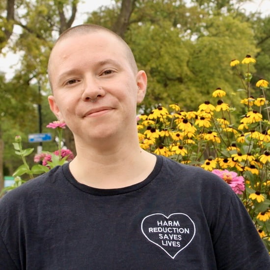 headshot of white trans-masculine human in front of flowers, wearing a black t-shirt that reads "Harm Reduction Saves Lives"